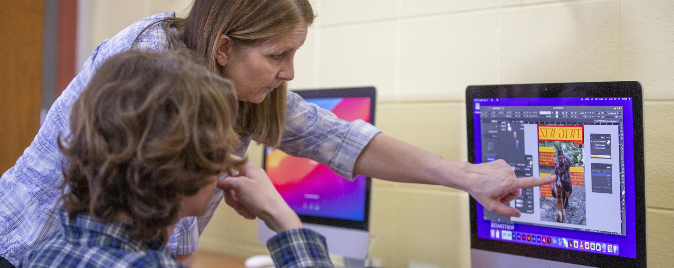 A faculty member points at a Mac computer as a student sits in a chair in front of the computer.