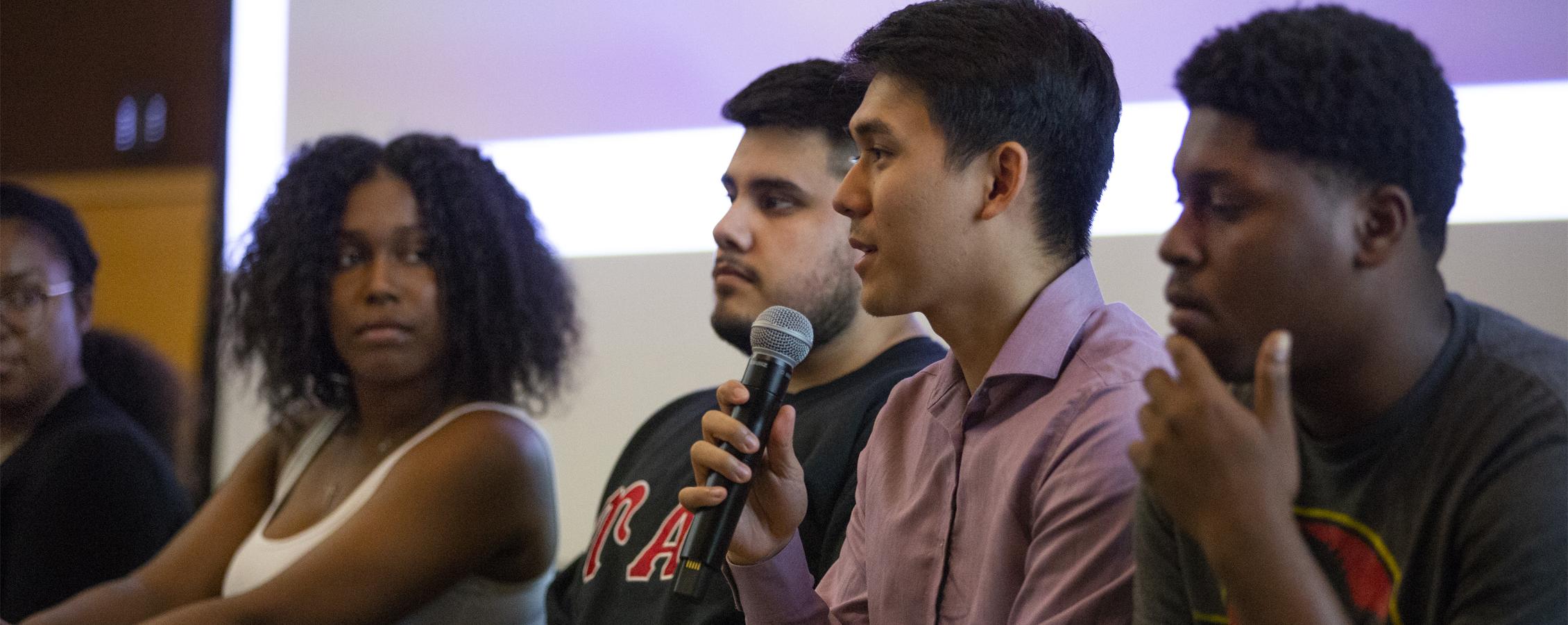 Four students are seated in the front of a room speaking into a microphone.