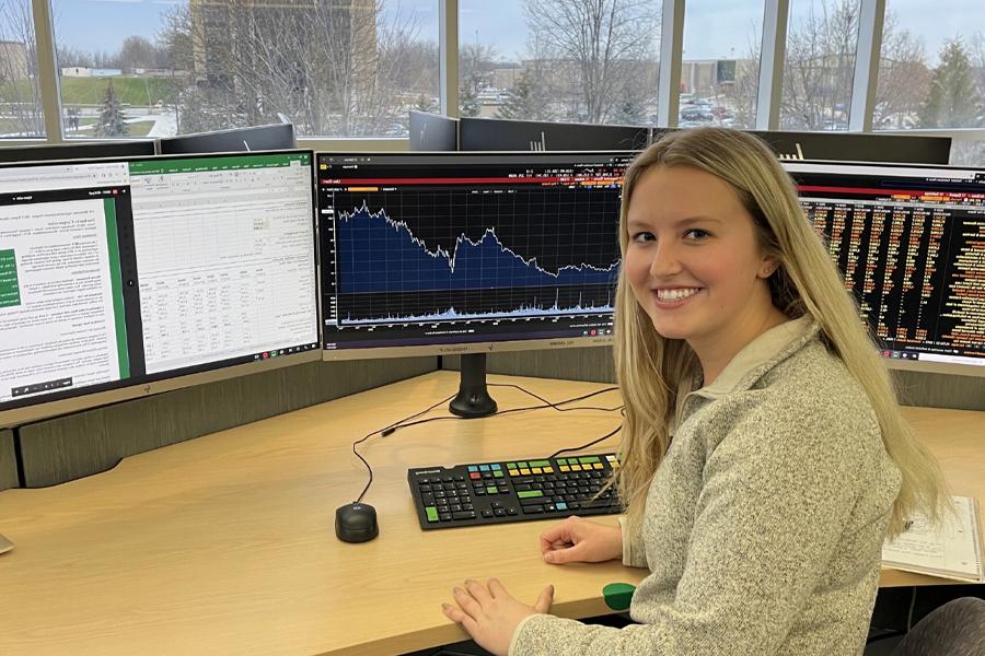 A student smiles at the camera and sits at a desk in front of several computer monitors.