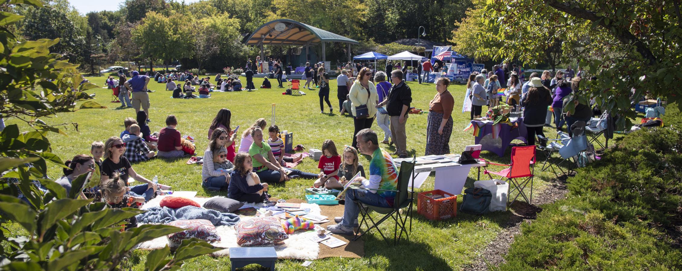 People gather in an outdoor green space surrounded by trees and pop up tents.