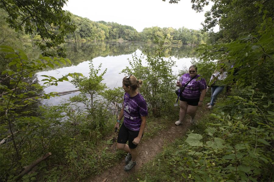 People walk along a trail through the woods and a clearing that shows a pond below.