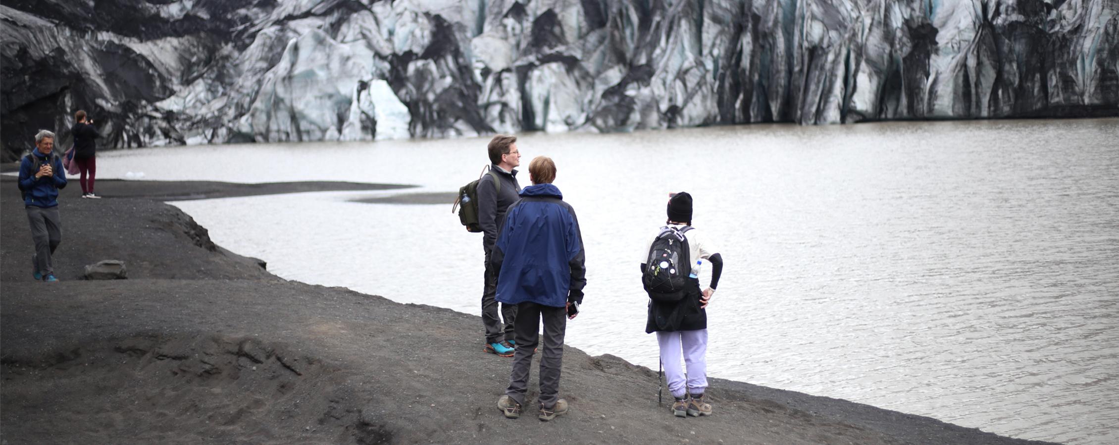 A faculty member stands with students on a shore with snow capped mountains in the background.